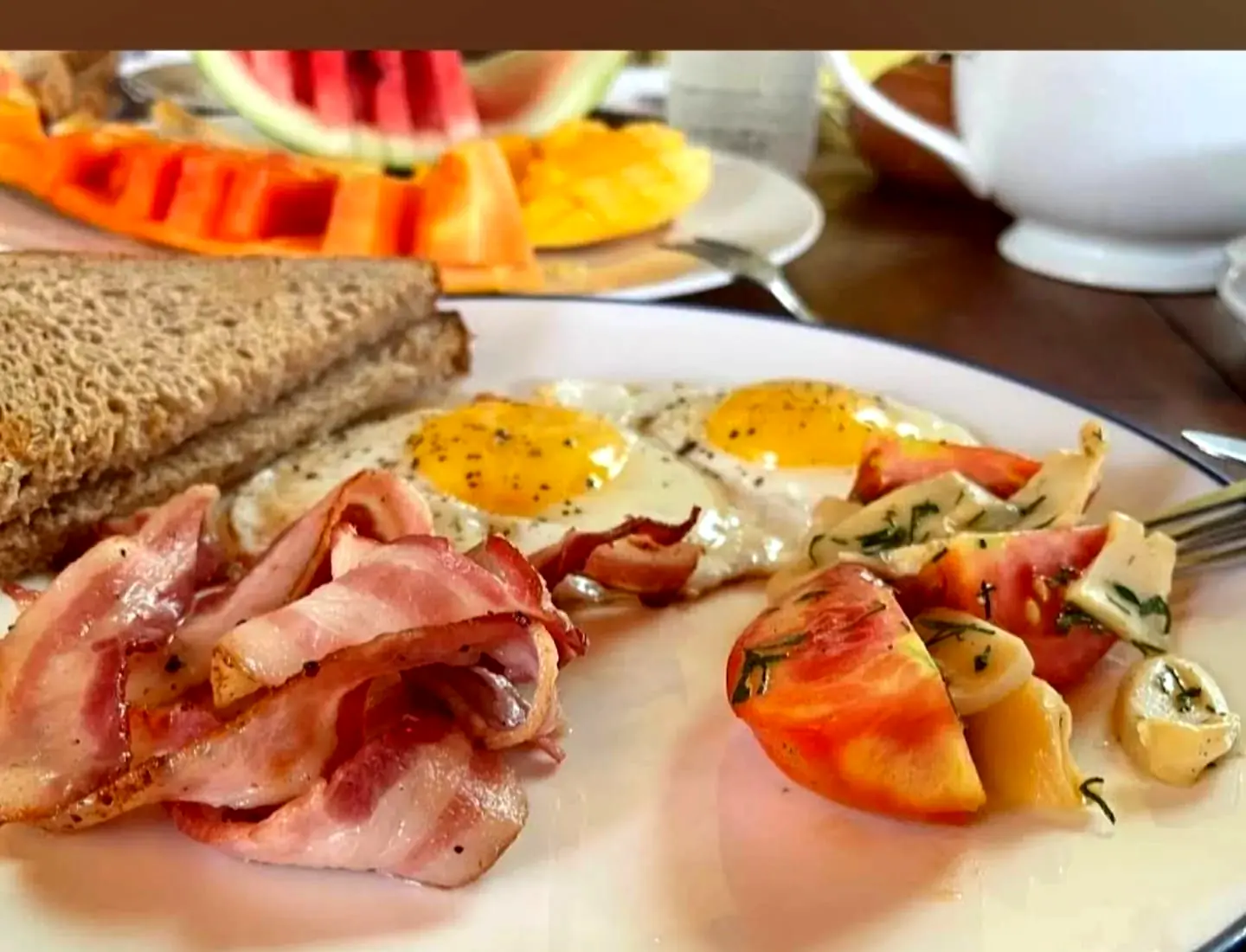 Breakfast plate with fried eggs, bacon, grilled tomato, toast and a side of fresh fruit