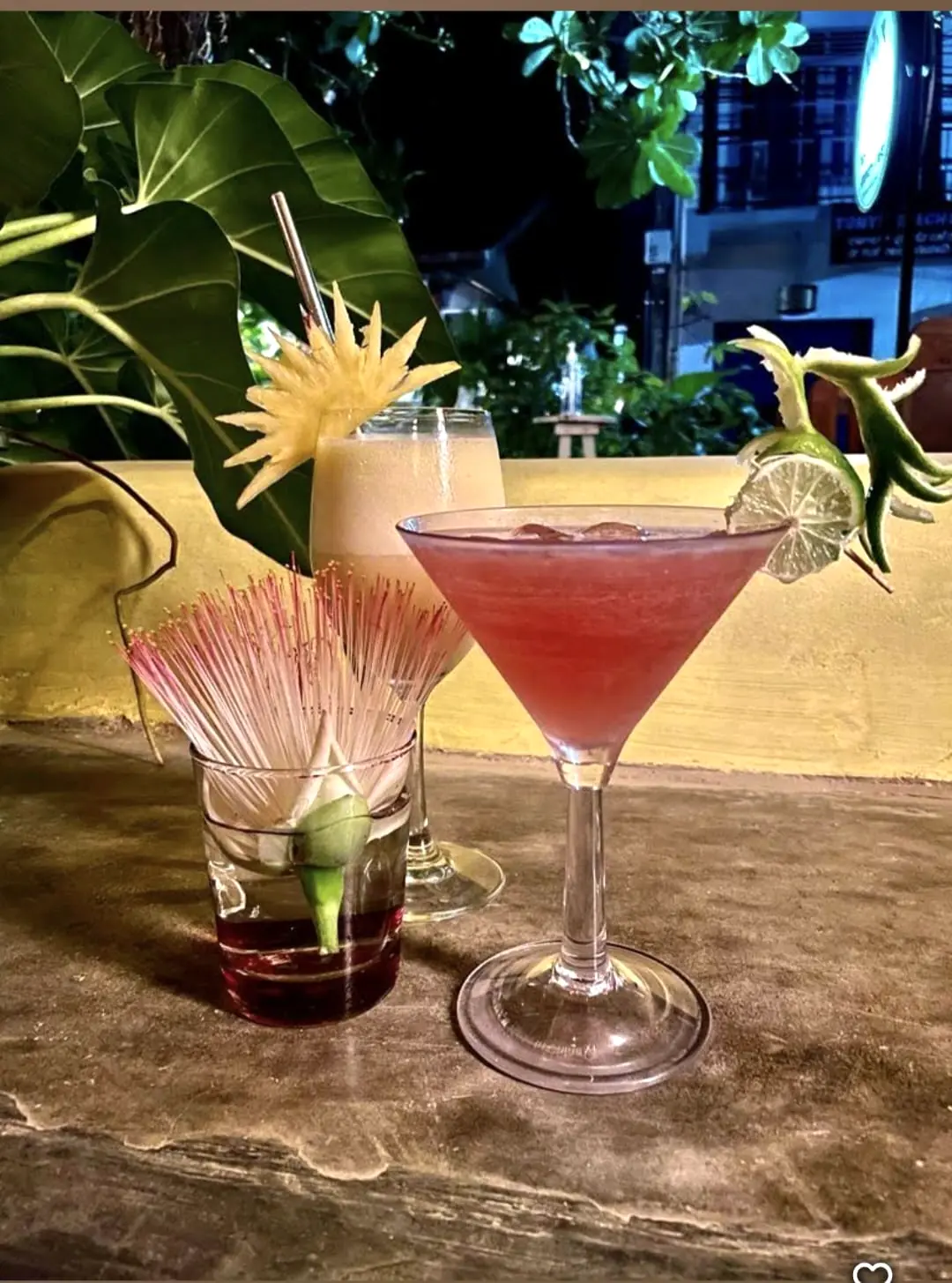 Two pink cocktails with fruit garnishes on a terrace table at night